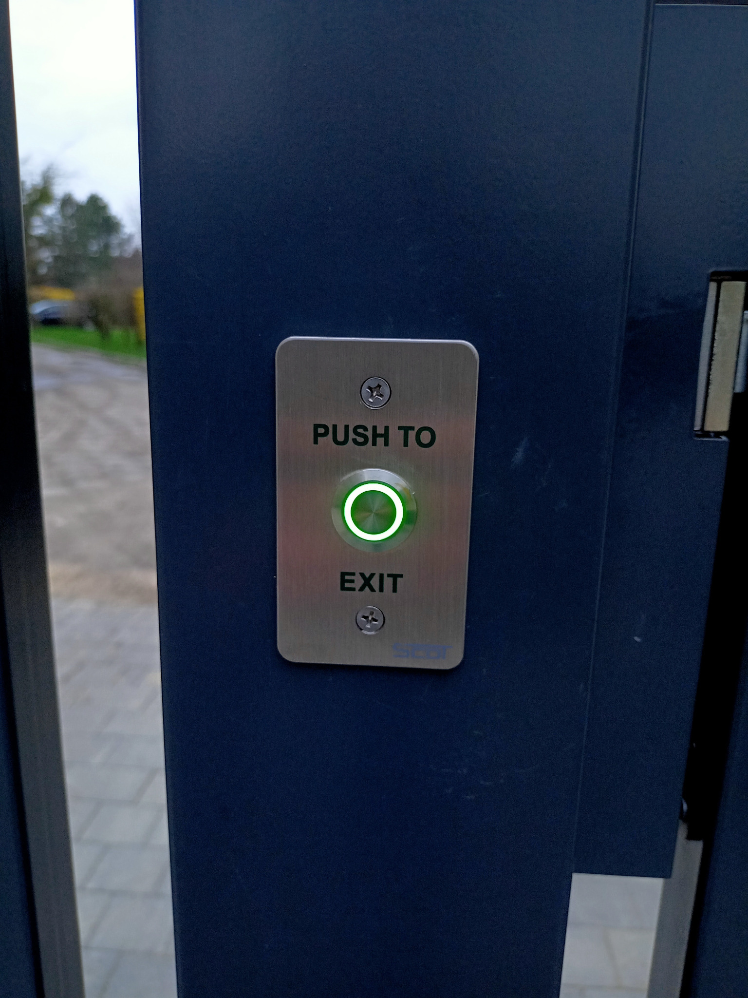 Przycisk otwierania bramy z napisem 'PUSH TO EXIT' i świecącym na zielono okręgiem, zamontowany na ciemnej, metalowej konstrukcji bramy.