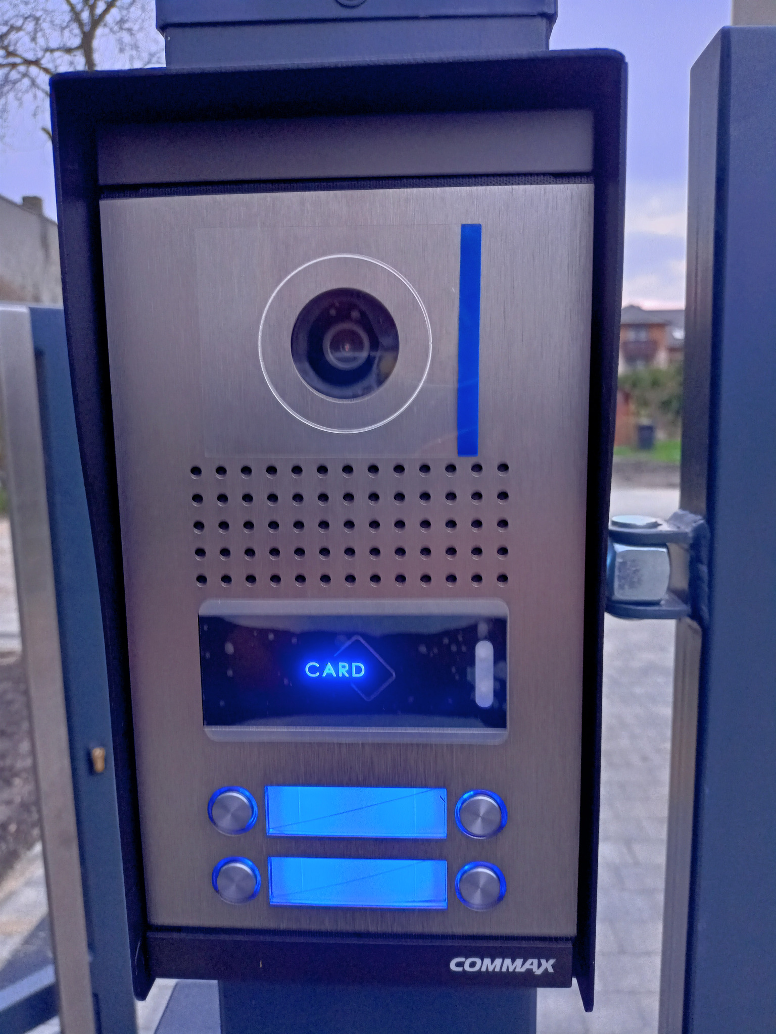 Zewnętrzny panel domofonowy Commax z kamerą, czytnikiem kart i podświetlanymi przyciskami, zamontowany na metalowym słupku.