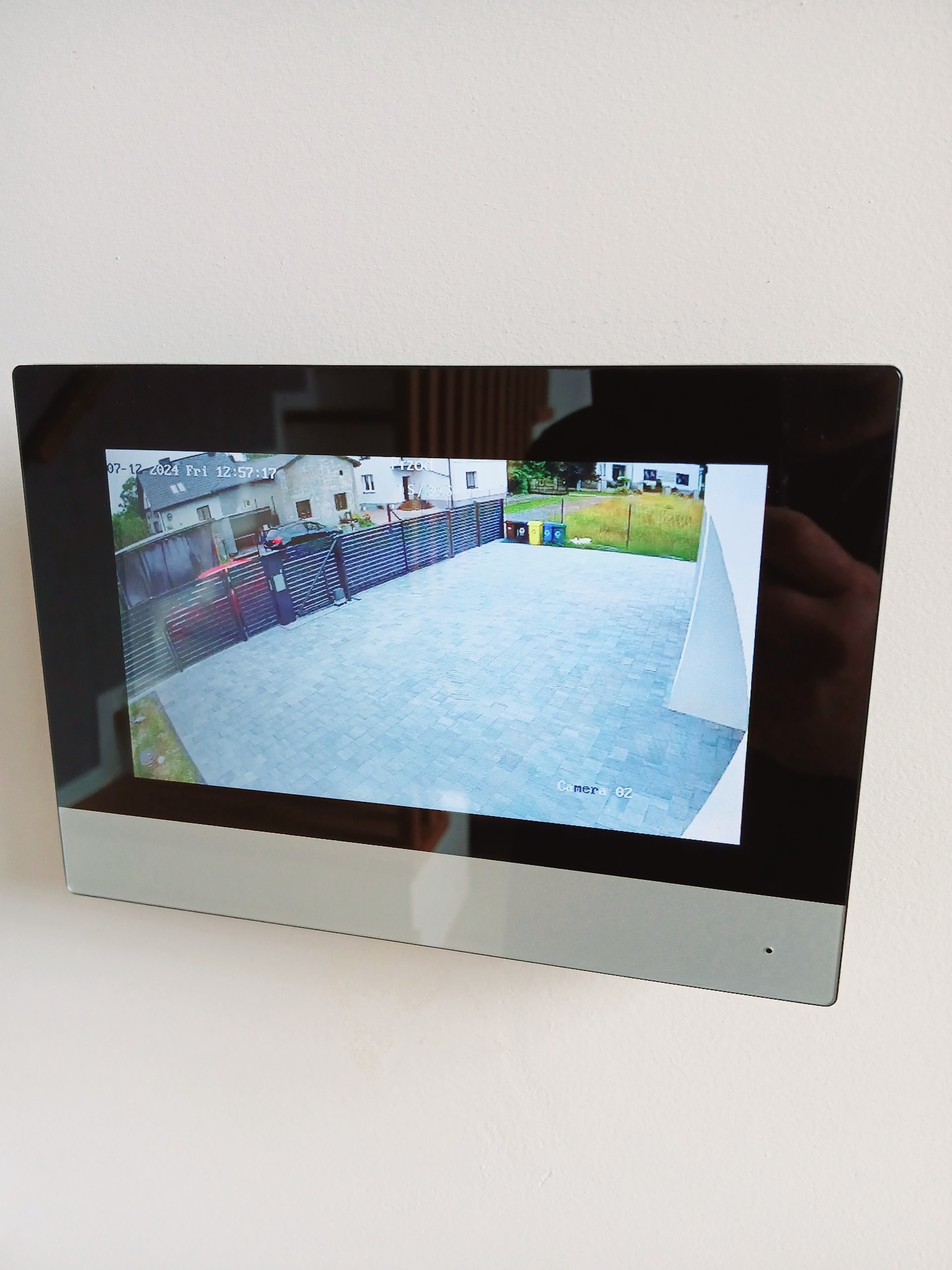 Ekran monitoringu domu z widokiem na podjazd, bramę i fragment ogrodu, data i godzina w lewym górnym rogu, kamera oznaczona jako 'camera 02'.