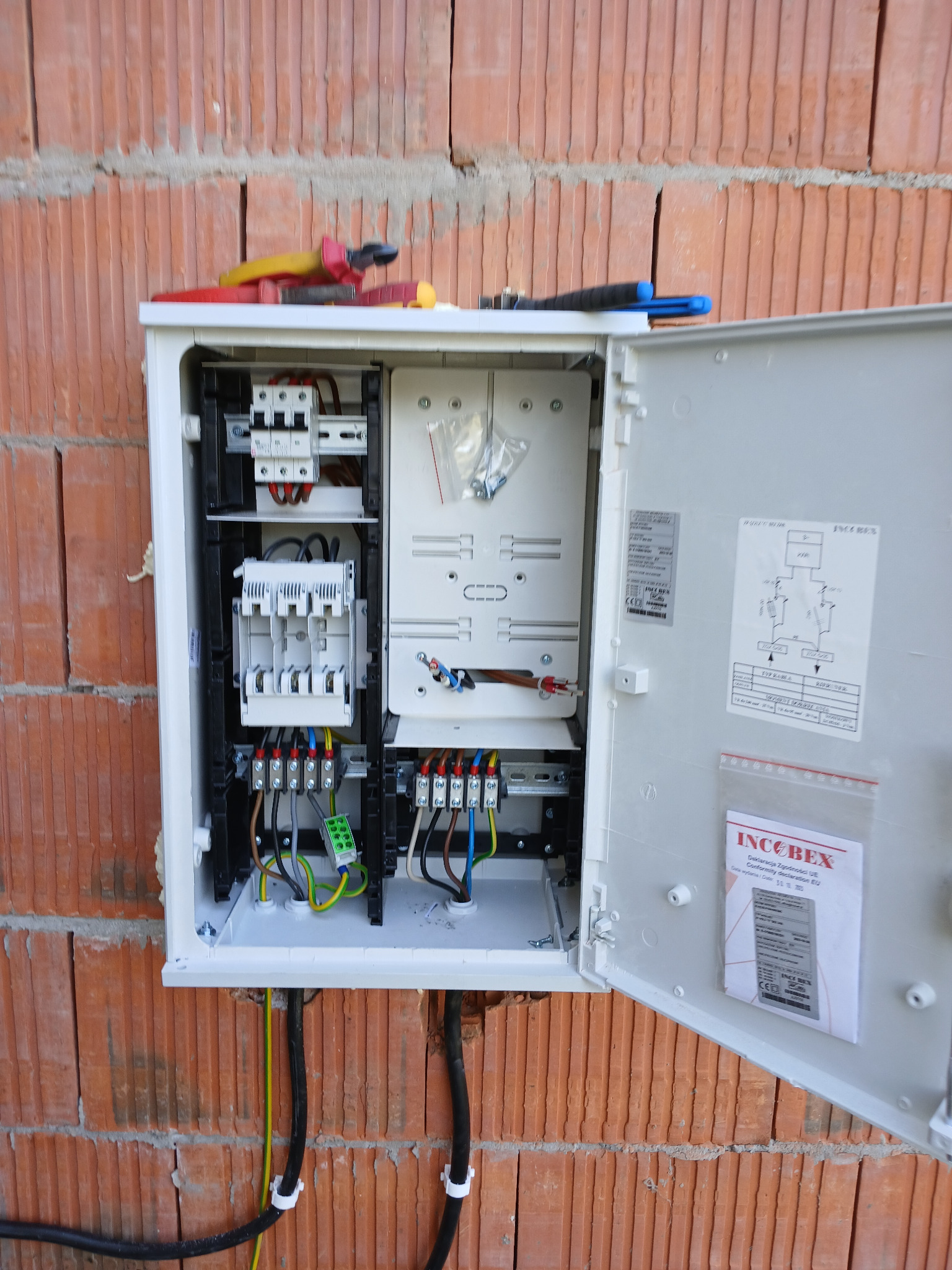 Otwarta skrzynka elektryczna z widocznymi bezpiecznikami, przewodami i schematem, zamontowana na ceglanej ścianie. Na górze skrzynki leżą kombinerki z żółto-czerwonymi rączkami. Widoczne czarne...