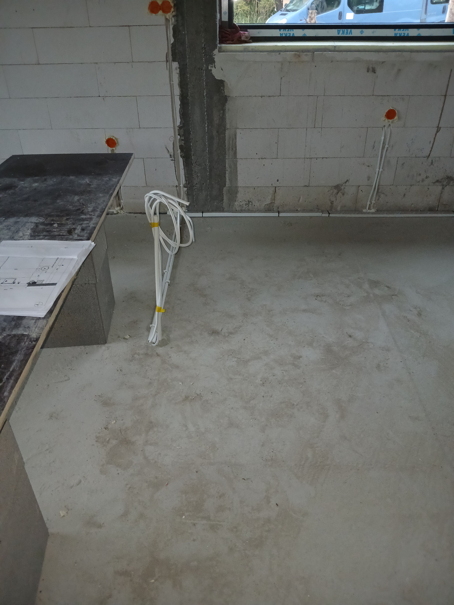 Surowy stan wnętrza z widocznymi elementami instalacji: przewody elektryczne zwinięte w pętlę, pomarańczowe puszki instalacyjne w ścianie z bloczków, częściowo wykończony filar i okno z widokiem...