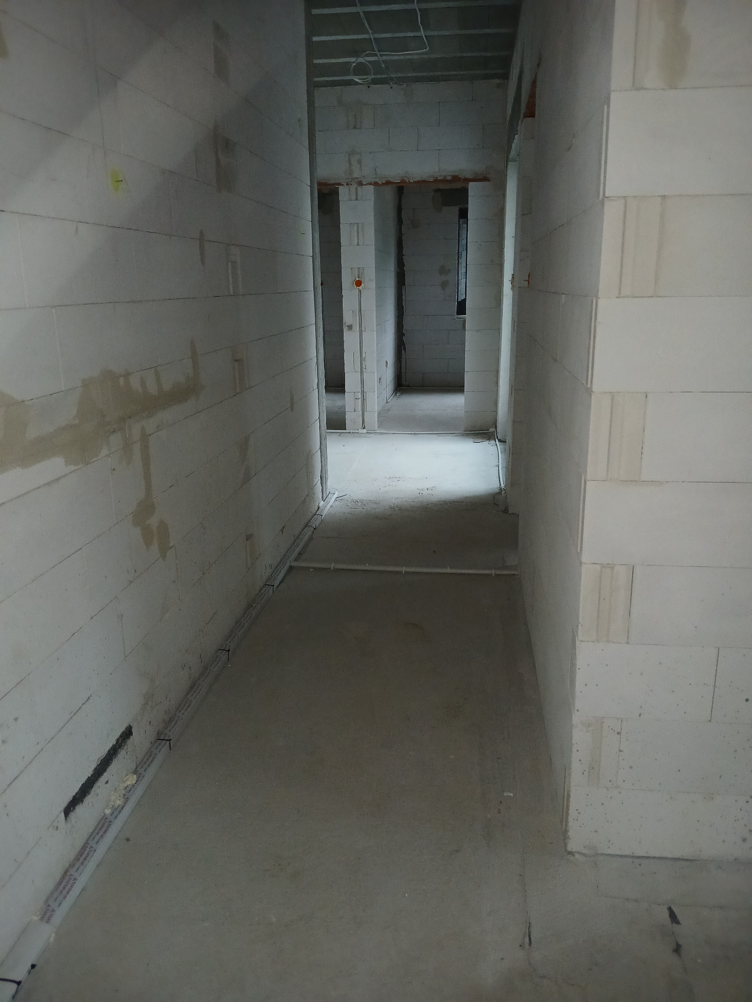 Korytarz w surowym stanie z widocznymi przewodami elektrycznymi w listwach przypodłogowych, ściany z betonu komórkowego, brak wykończenia.