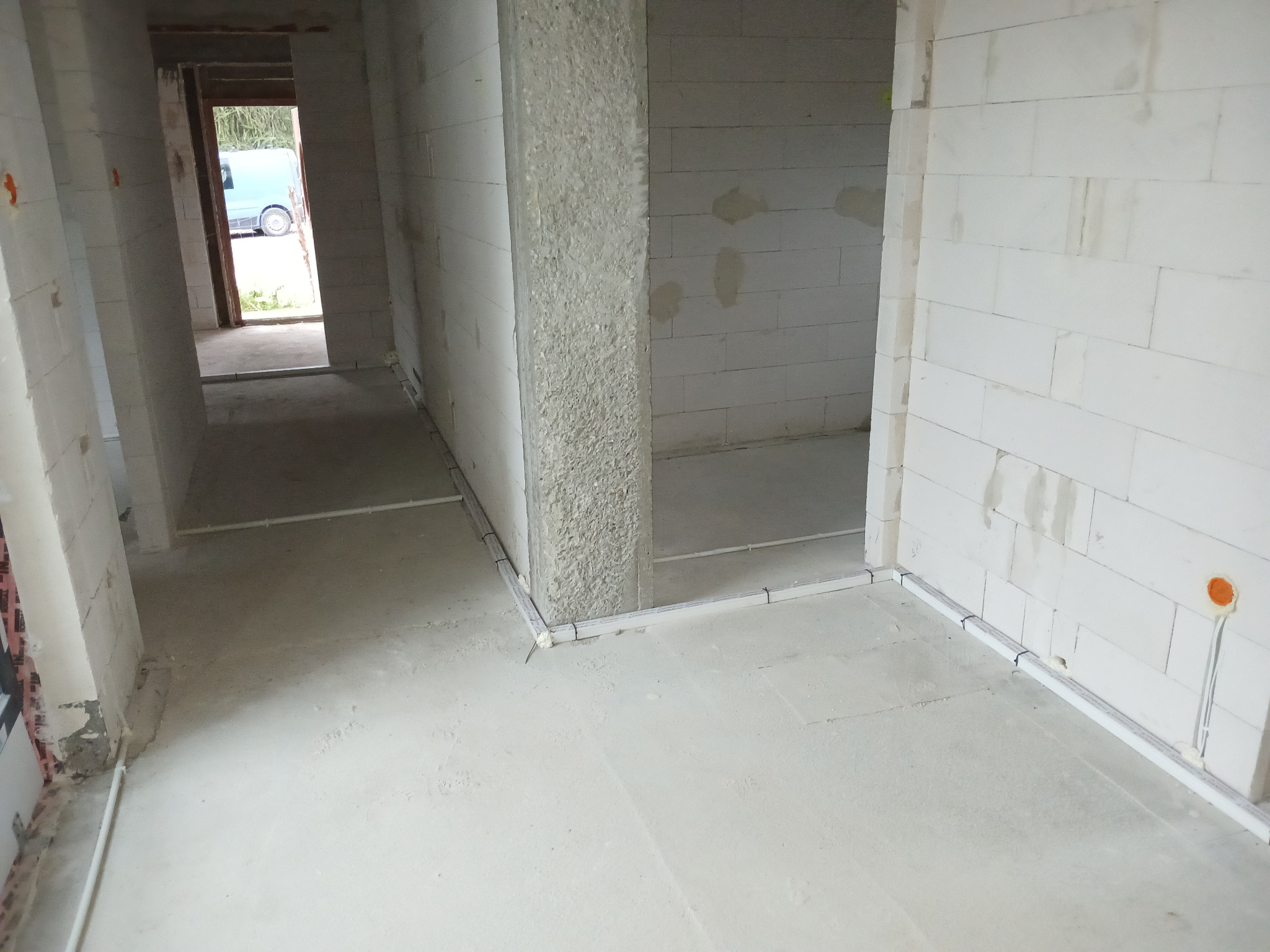 Surowy stan wnętrza budynku w trakcie prac instalacyjnych, widoczne białe ściany z betonu komórkowego, szare wylewki podłogowe oraz rury instalacyjne prowadzone wzdłuż ścian z widocznymi punktami...