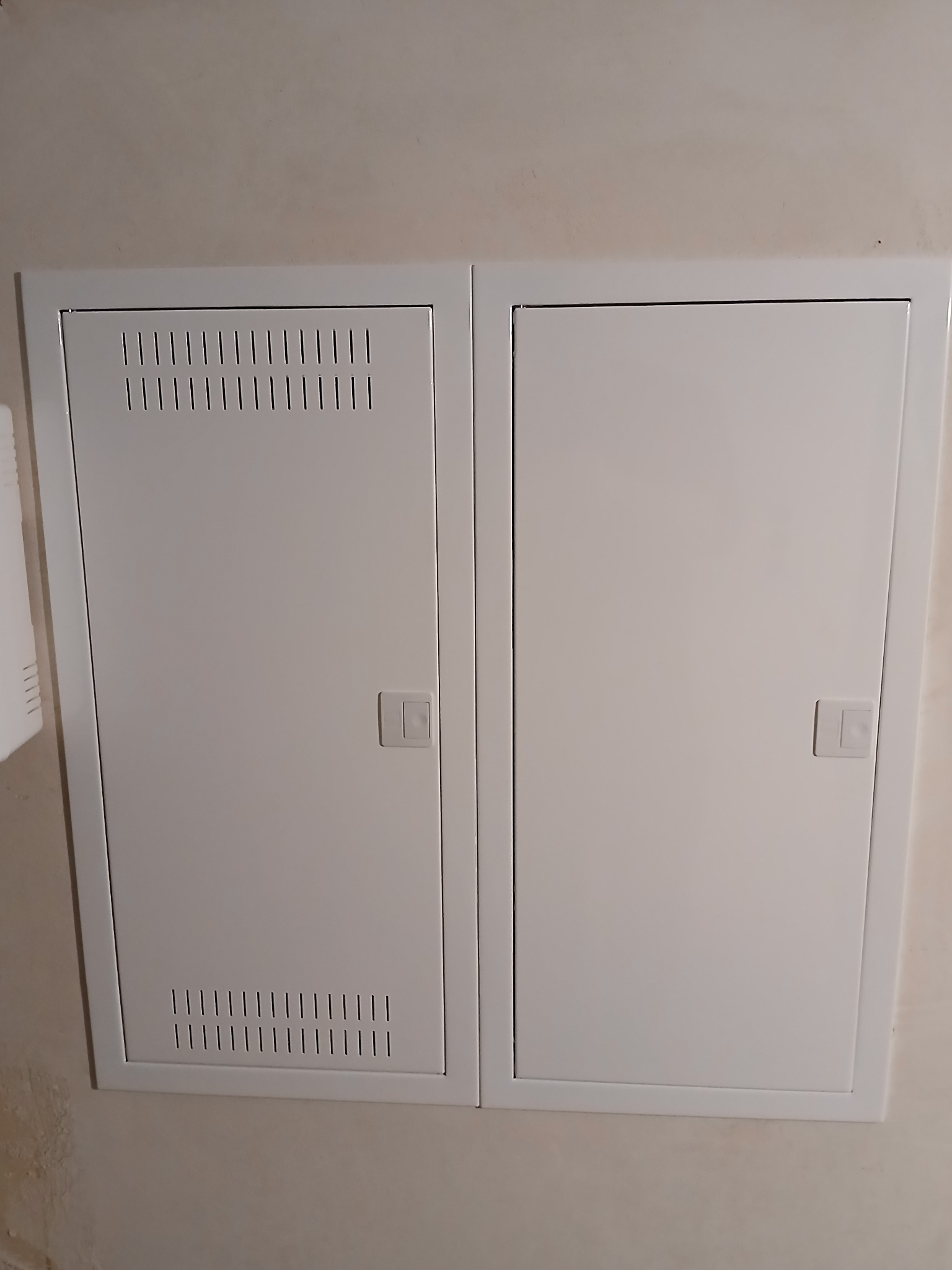 Biała, podwójna skrzynka elektryczna z wentylacją na jednej z klap, zamontowana na ścianie wewnątrz budynku.