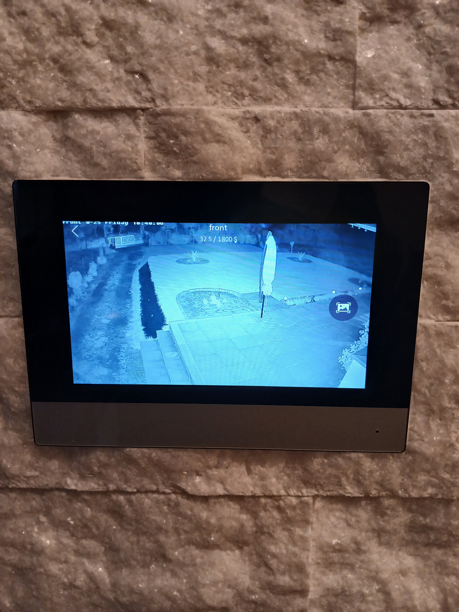 Ekran monitoringu domu zamontowany na ścianie z widokiem na podjazd i ogród w nocy, z datą i godziną w lewym górnym rogu ekranu.