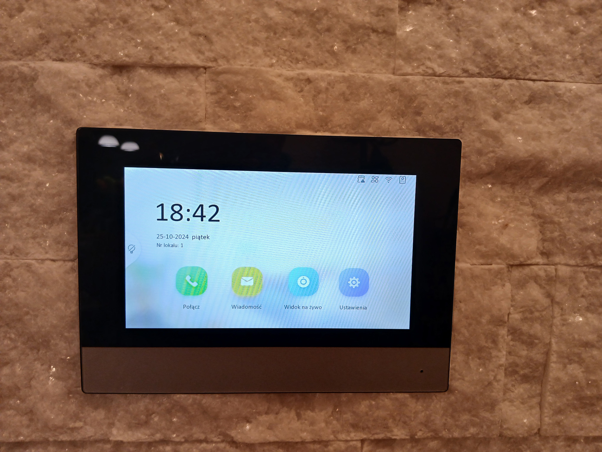 Panel dotykowy wideodomofonu zamontowany na ścianie z dekoracyjnej cegły, wyświetlający godzinę, datę i ikony funkcji: połączenie, wiadomość, podgląd na żywo i ustawienia.