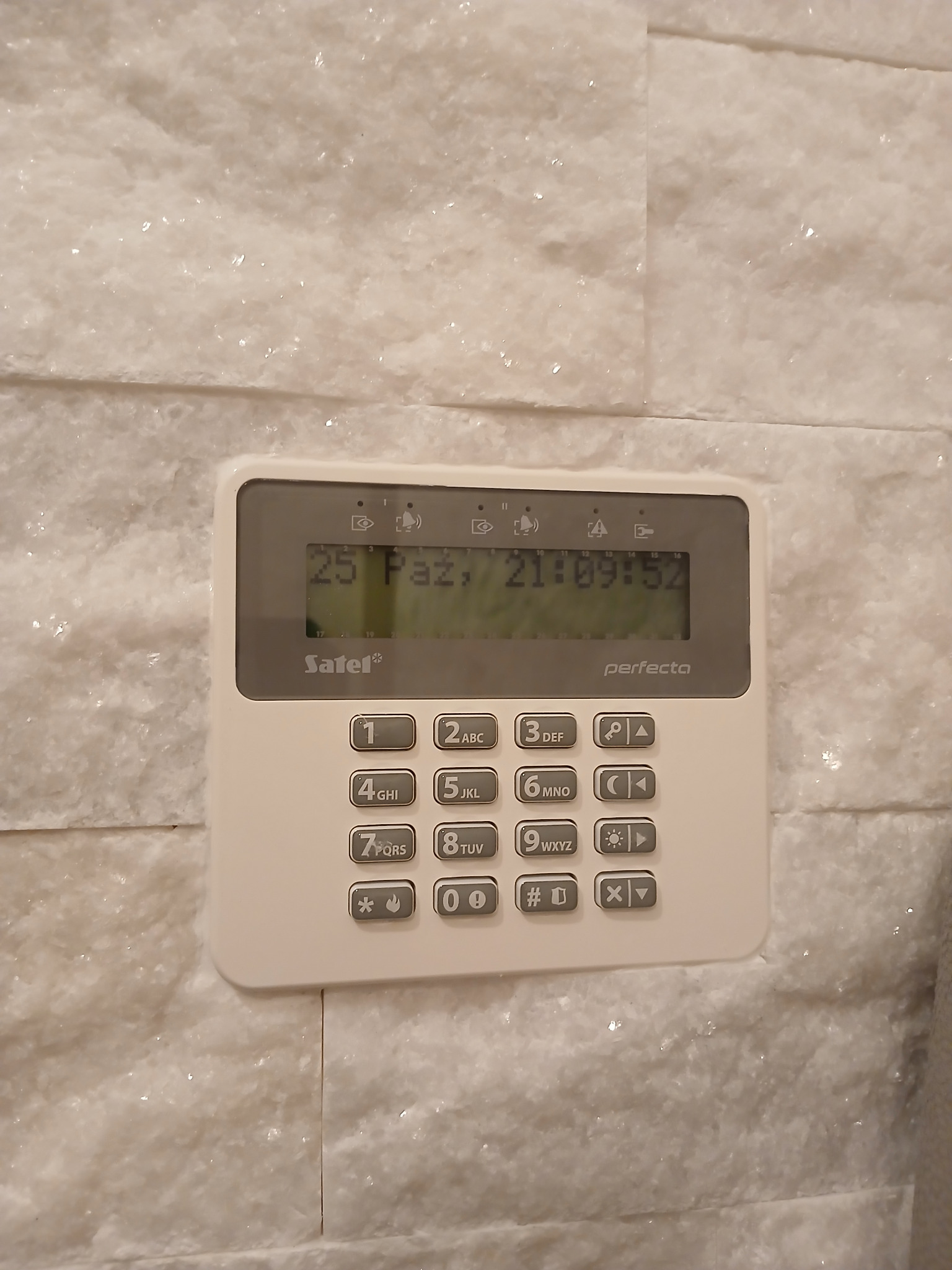 Panel sterowania alarmem Satel Perfecta zamontowany na ścianie z dekoracyjnej białej cegły, wyświetlający datę i godzinę.