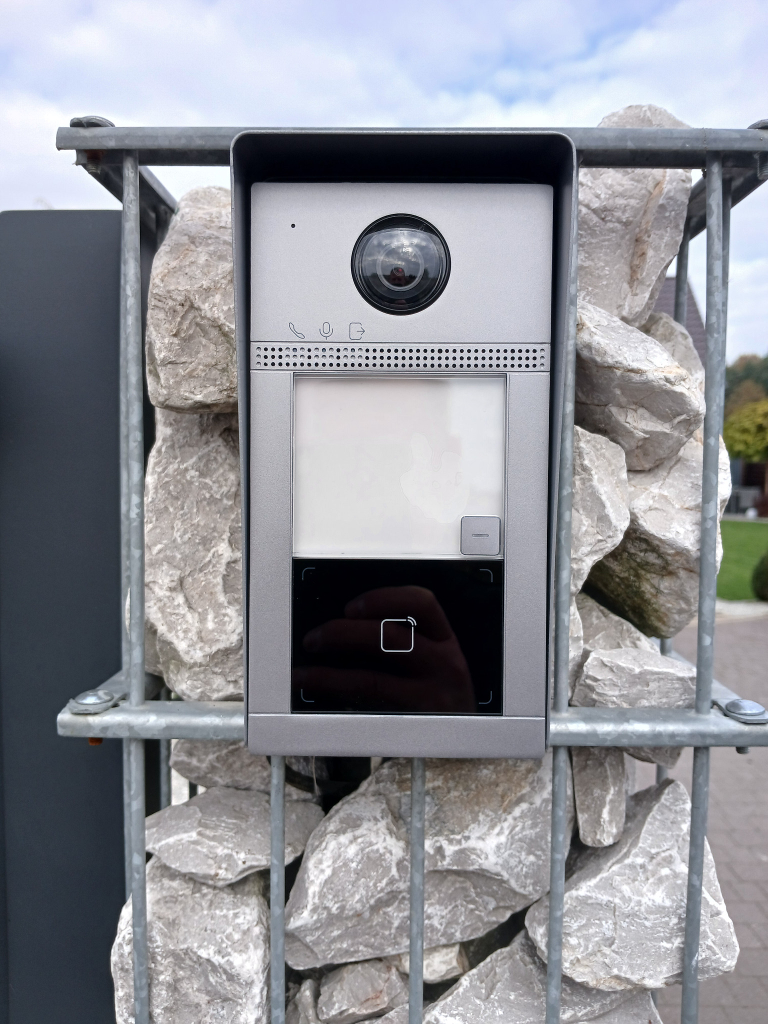 Szary wideodomofon z kamerą zamontowany na ogrodzeniu z kamieni w metalowej siatce, widoczny fragment chodnika i zieleni w tle.