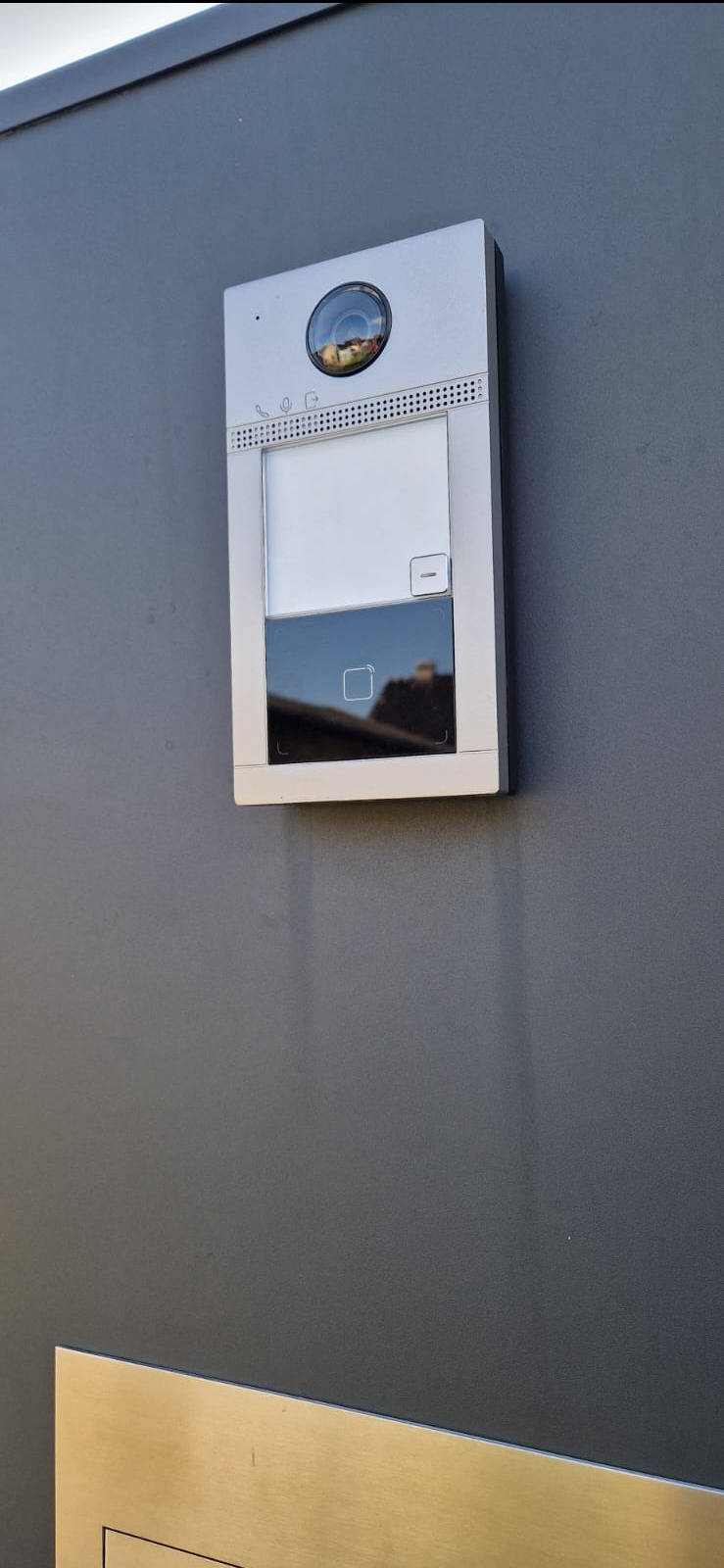 Srebrny wideodomofon z kamerą i dotykowym panelem, zamontowany na szarej ścianie zewnętrznej budynku. W obiektywie kamery widoczne odbicie otoczenia.