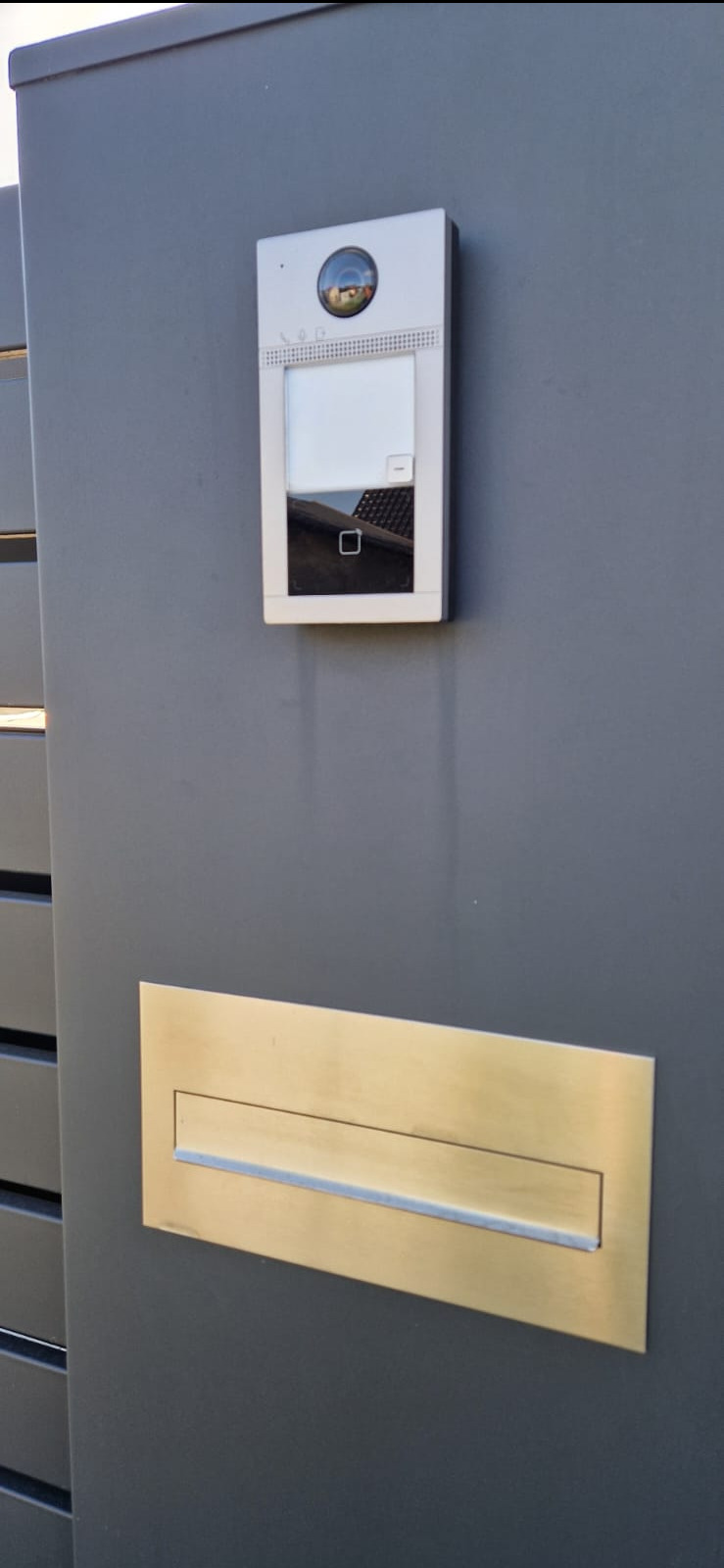 Szary panel ogrodzeniowy z zamontowanym wideodomofonem z kamerą i złotą skrzynką na listy