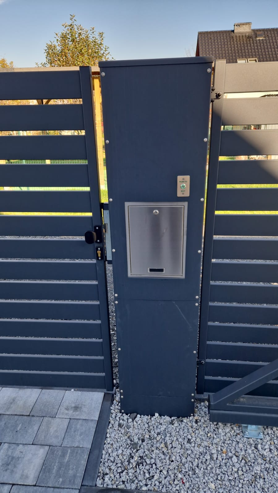 Nowoczesna, grafitowa brama wjazdowa z poziomymi panelami, wyposażona w skrzynkę na listy ze stali nierdzewnej i przycisk dzwonka, osadzona na żwirowym podłożu.