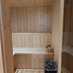 Wnętrze sauny z jasnego drewna, z piecem na kamienie i drewnianym wiadrem z chochlą na ławce.