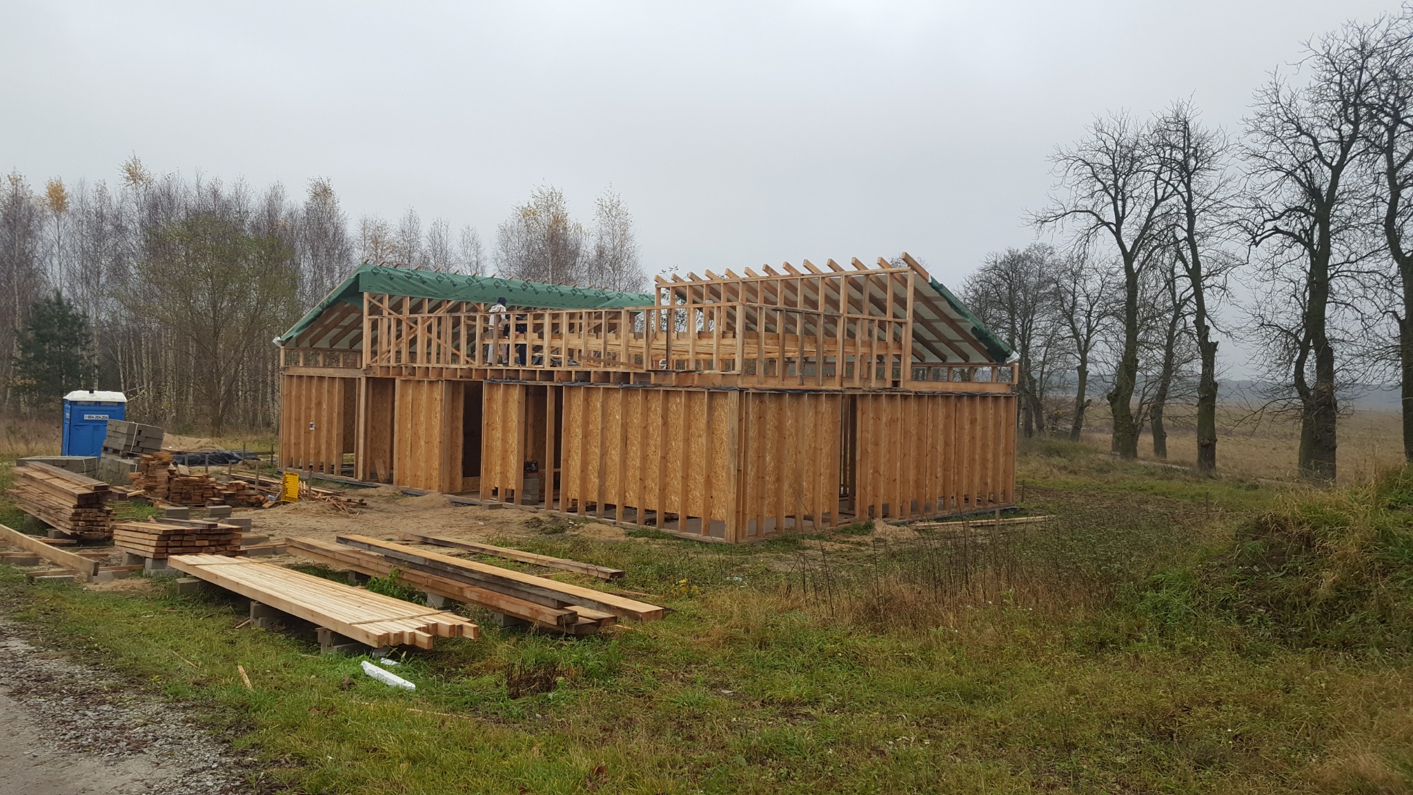 Wznoszenie konstrukcji szkieletowej domu z widocznym zielonym pokryciem dachu i stertami drewna na zewnątrz, na tle jesiennego krajobrazu.