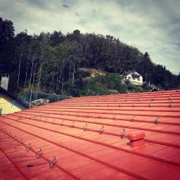 Fox soluction - Nowy, czerwony dach z ceramicznej dachówki z zamontowanymi hakami przeciwśniegowymi, widok na zalesione wzgórze i dom w tle.