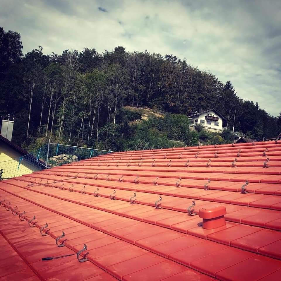 Nowy, czerwony dach z ceramicznej dachówki z zamontowanymi hakami przeciwśniegowymi, widok na zalesione wzgórze i dom w tle.