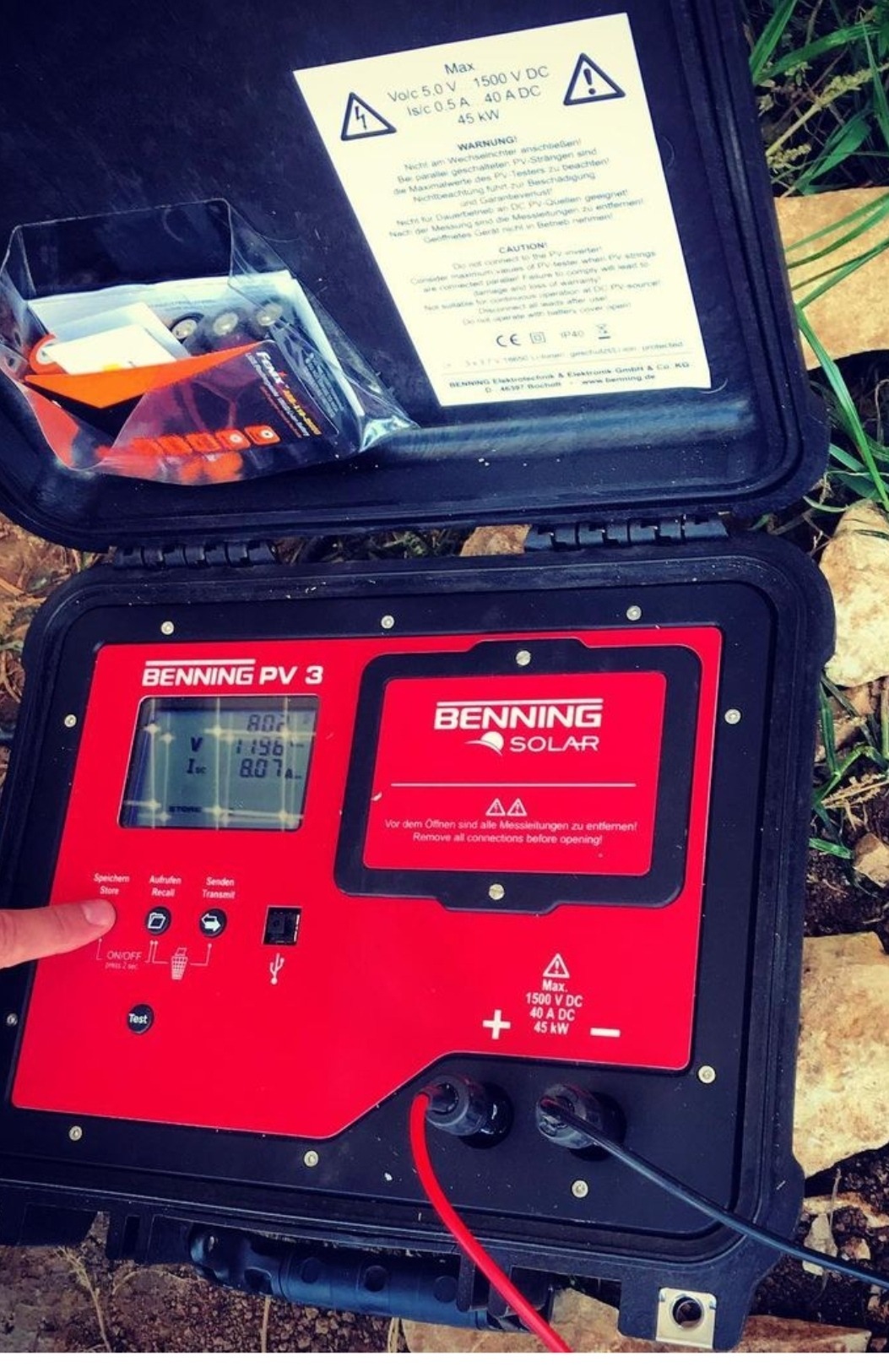 Otwarta walizka z miernikiem Benning PV 3 do pomiaru parametrów instalacji fotowoltaicznych, widoczne podłączone przewody pomiarowe, wyświetlacz z odczytami, instrukcja wewnątrz wieka i akcesoria...