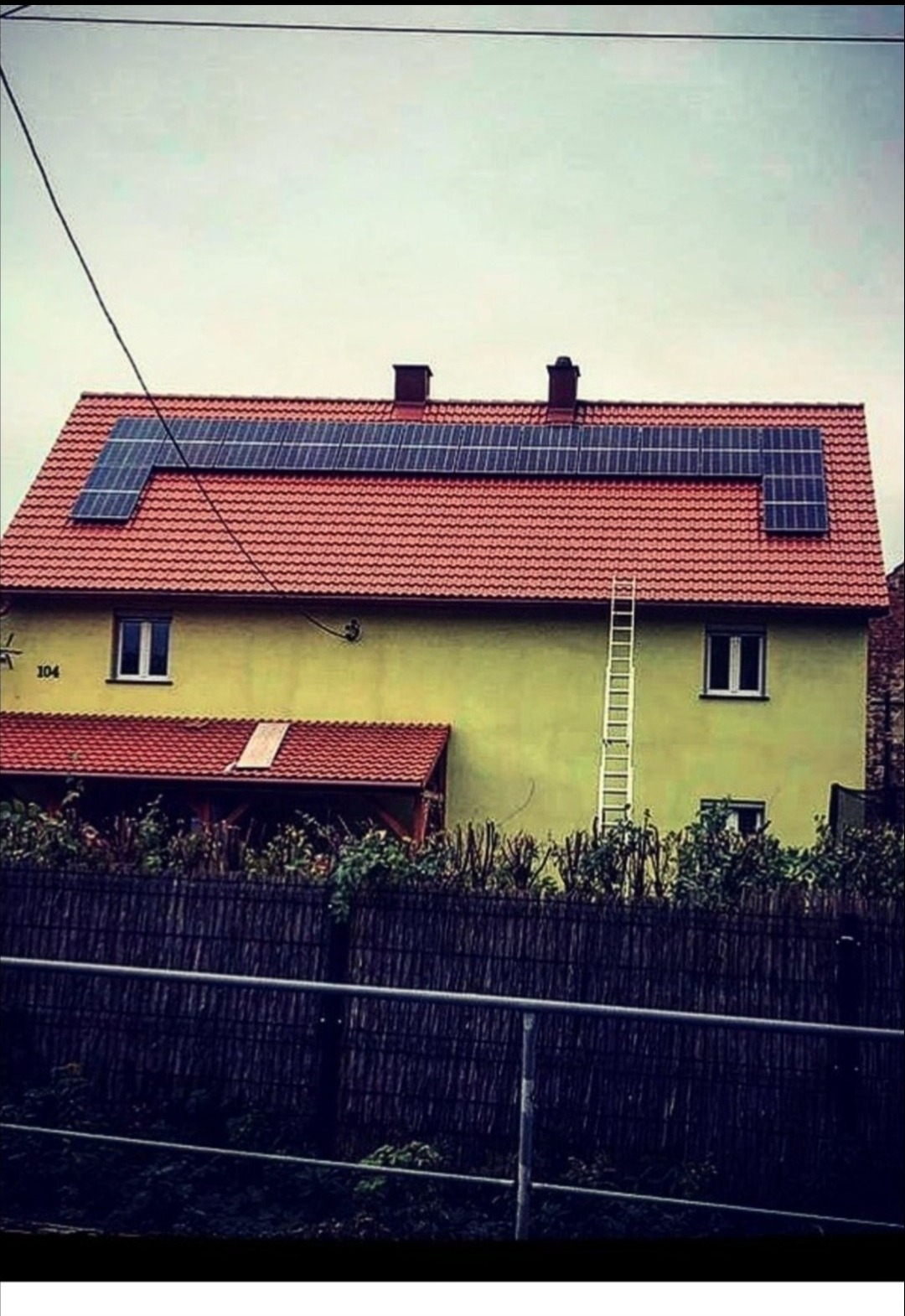 Dom jednorodzinny z panelami słonecznymi na czerwonym dachu, drabina oparta o ścianę, widok zza ogrodzenia.