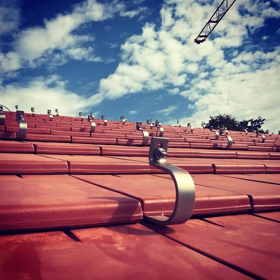 Czerwona dachówka z zamontowanymi metalowymi uchwytami pod instalację, widok z dołu na błękitne niebo z chmurami i żuraw budowlany w oddali.