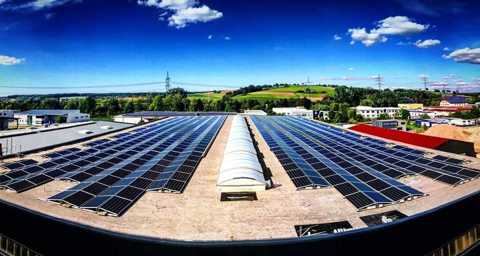 Rozległa instalacja paneli słonecznych na dachu budynku przemysłowego, widok z góry, błękitne niebo z chmurami w tle.