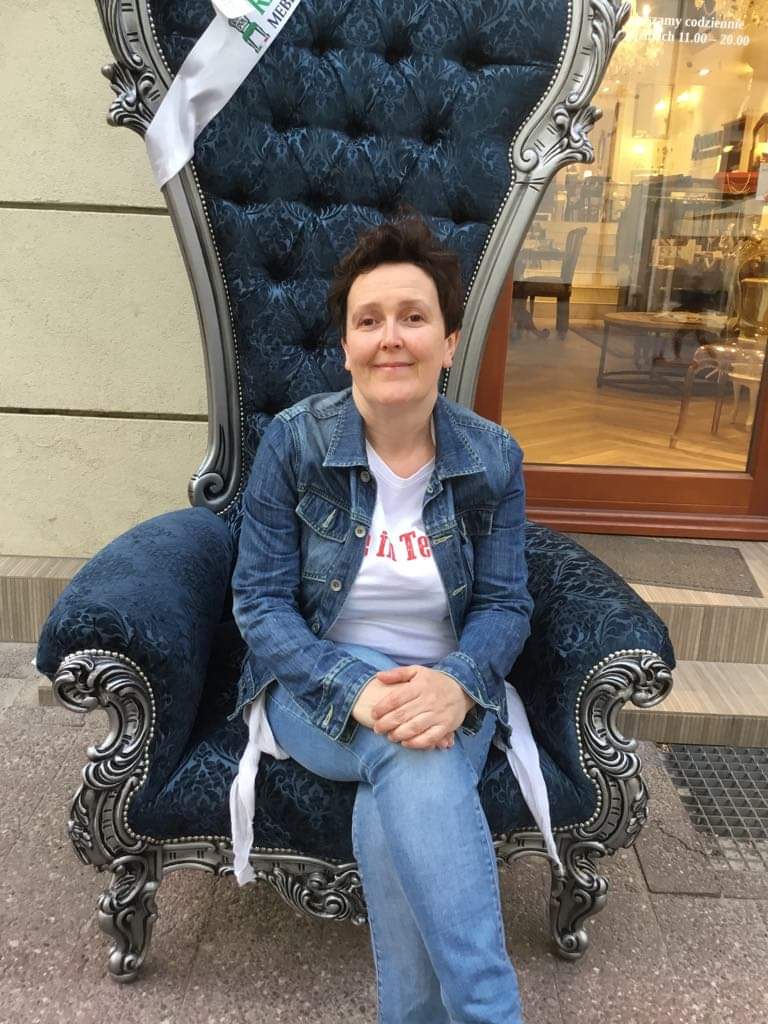 Kobieta siedzi na bogato zdobionym, niebieskim fotelu w stylu królewskim przed witryną sklepu meblowego. Fotel ma srebrne zdobienia i wysokie oparcie.