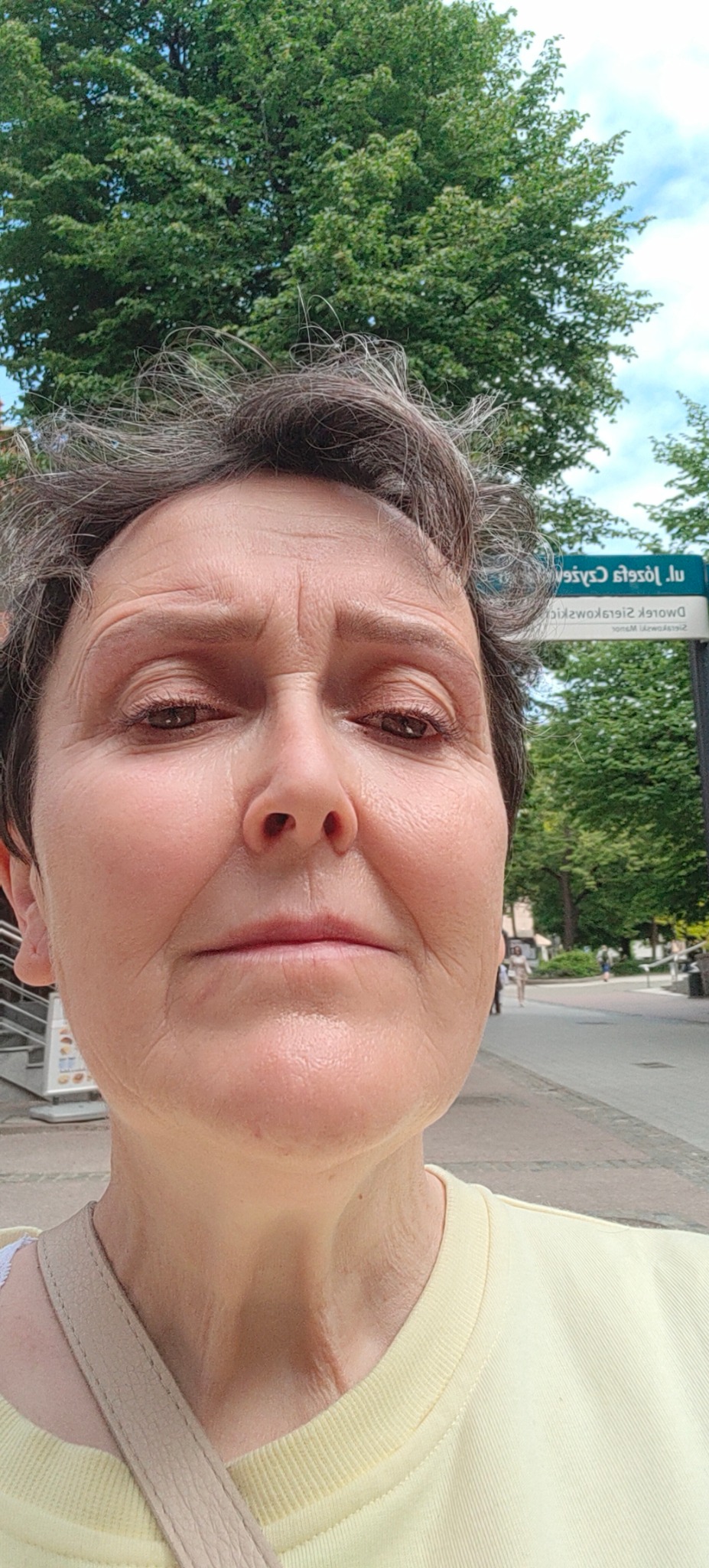 Portret kobiety z bliska na tle zieleni miejskiej i tabliczki z nazwą ulicy. Widoczne zmarszczki i naturalny wygląd.