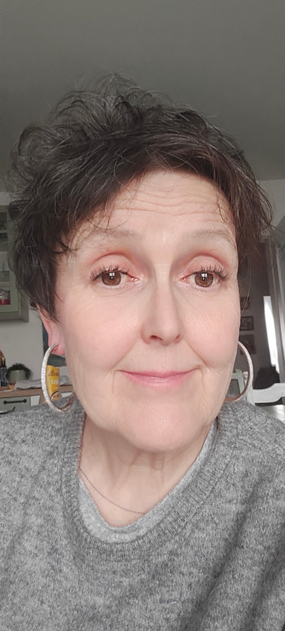 Portret kobiety z brązowymi oczami i srebrnymi kolczykami kołami, uśmiechająca się, w szarym swetrze, z krótkimi ciemnymi włosami.