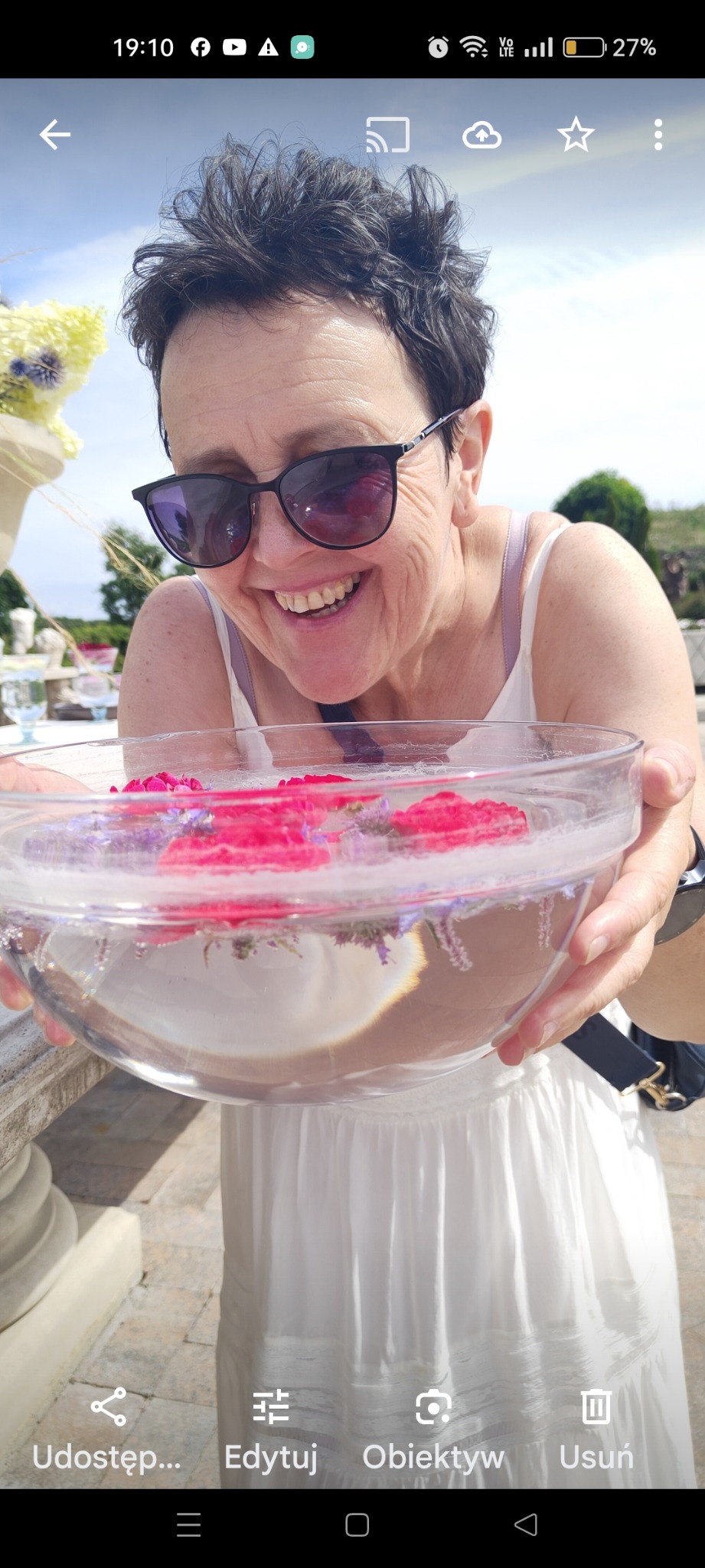 Uśmiechnięta kobieta w okularach prezentuje szklaną misę z kwiatami w wodzie na tle jasnego otoczenia, prawdopodobnie dekoracja lub element ceremonii.