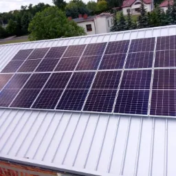 Panel słoneczny zamontowany na blaszanym dachu z widocznymi przewodami, w tle zabudowa jednorodzinna i drzewa.