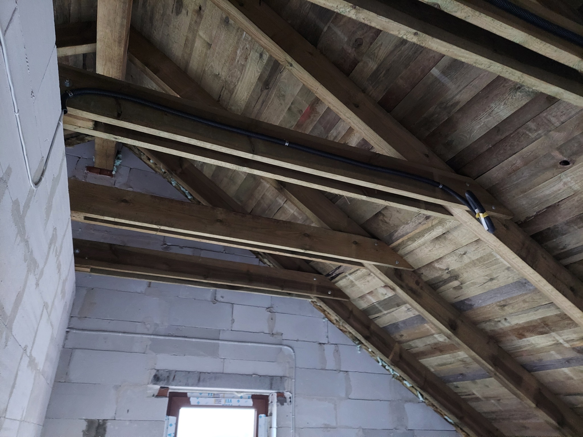 Czarna rura karbowana z kablami elektrycznymi zamocowana do drewnianej konstrukcji dachu na poddaszu, widoczne ściany z białych bloczków betonowych.