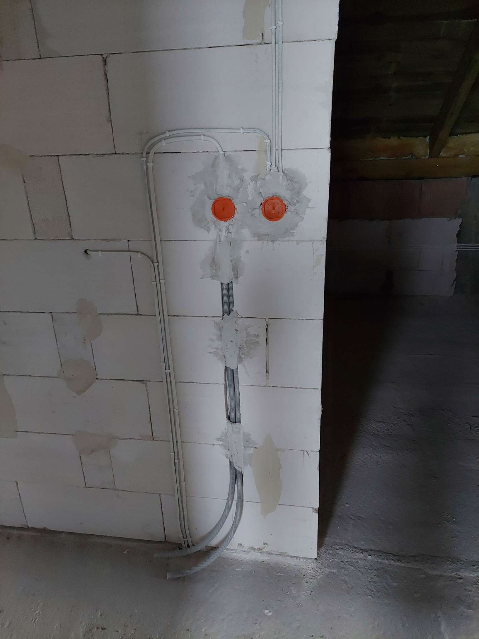 Świeżo położone kable elektryczne w peszlach i rurkach mocowane do ściany z betonu komórkowego, widoczne pomarańczowe puszki instalacyjne, częściowo zamurowane.