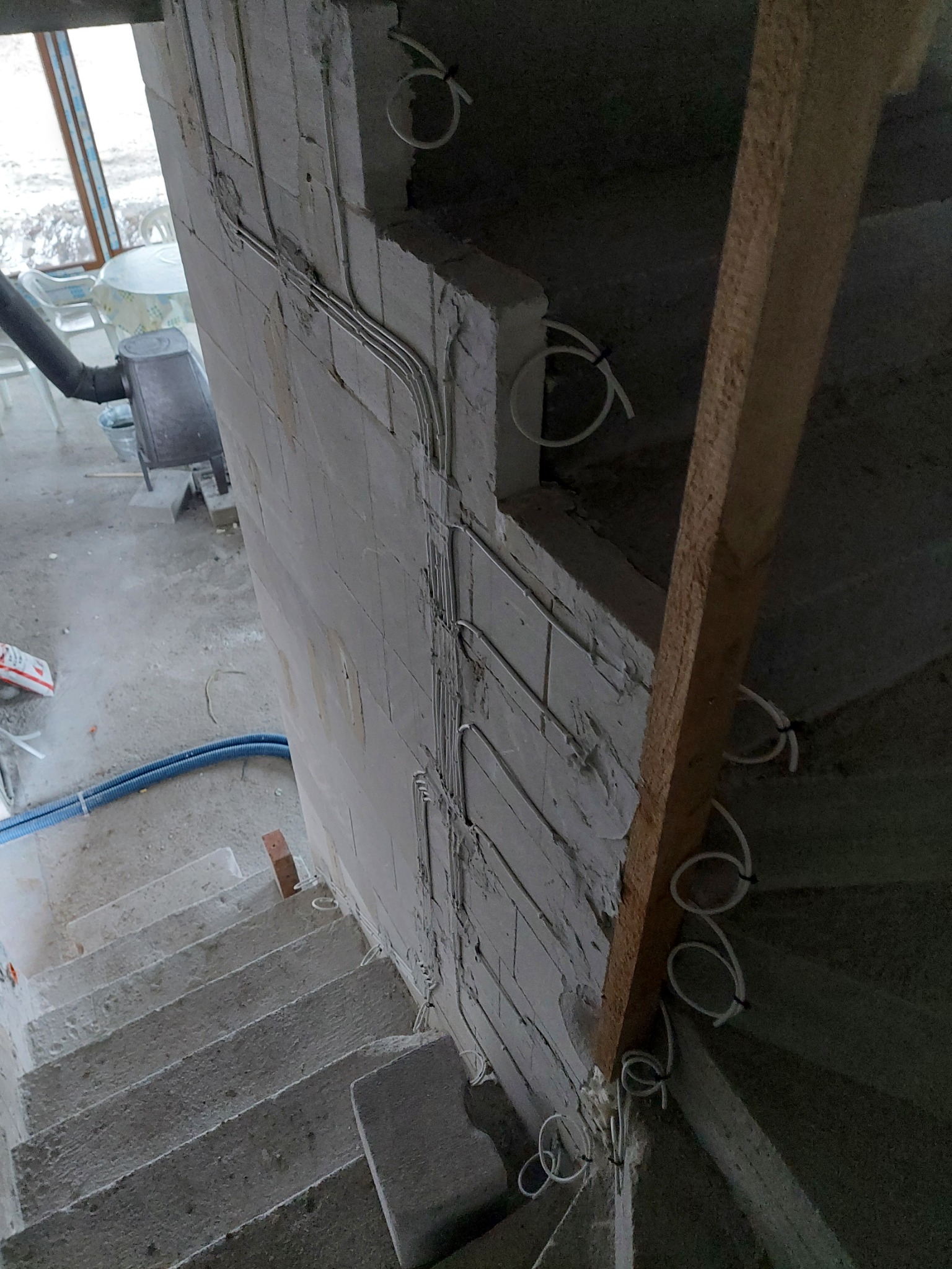 Wewnętrzna ściana klatki schodowej w trakcie prac instalacyjnych, widoczne kable elektryczne poprowadzone w peszlach i przymocowane do ściany z betonu komórkowego, w tle piec grzewczy.