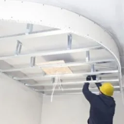 Montaż metalowej konstrukcji sufitu podwieszanego z płyt kartonowo-gipsowych, widoczne profile, mocowania i fragment płyty, pracownik w kasku. W tle białe ściany i fragment istniejącego sufitu.