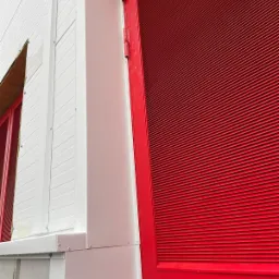 Fragment ściany hali z białych płyt warstwowych z widocznym oknem i czerwonymi drzwiami z poziomymi żaluzjami, uchwycony pod kątem z bliska.