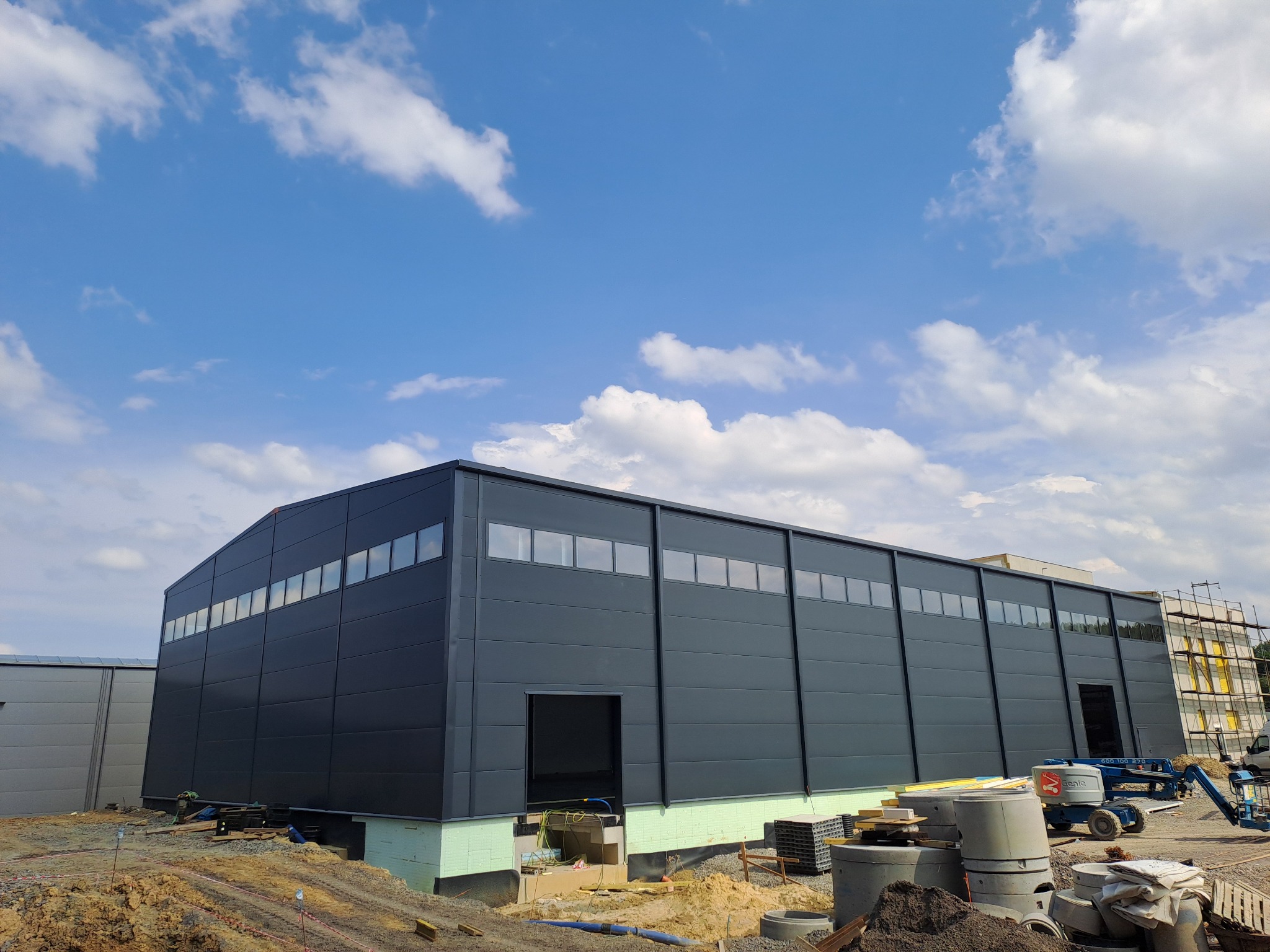 Nowoczesna hala przemysłowa w trakcie budowy, z widocznymi panelami warstwowymi, fundamentami i sprzętem budowlanym na placu budowy, na tle błękitnego nieba z obłokami.