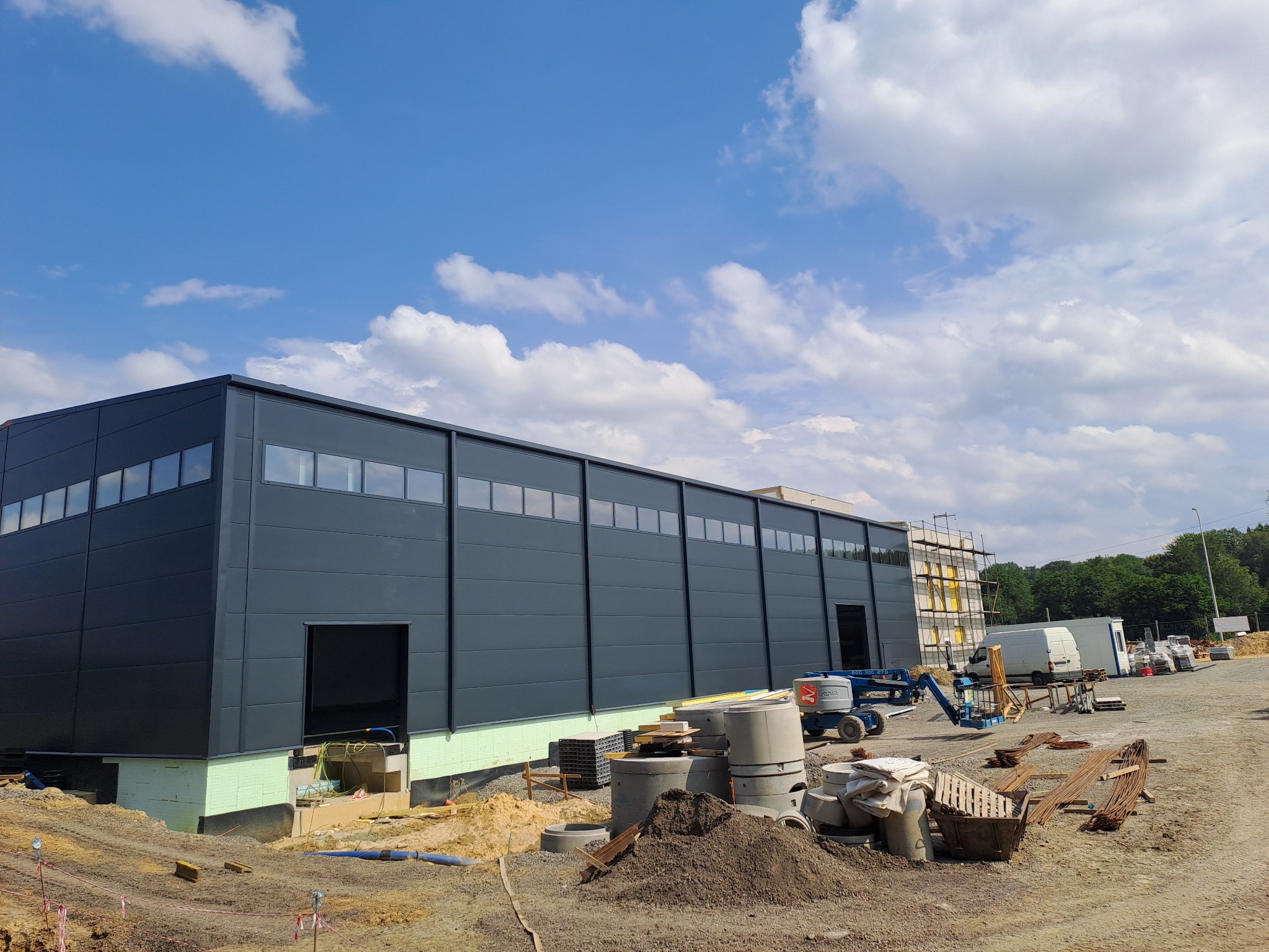 Nowoczesna hala w trakcie budowy, widoczna szara elewacja z oknami, fundamenty ocieplone styropianem, na placu budowy leżą betonowe kręgi, w tle podnośnik koszowy i zaparkowany bus.