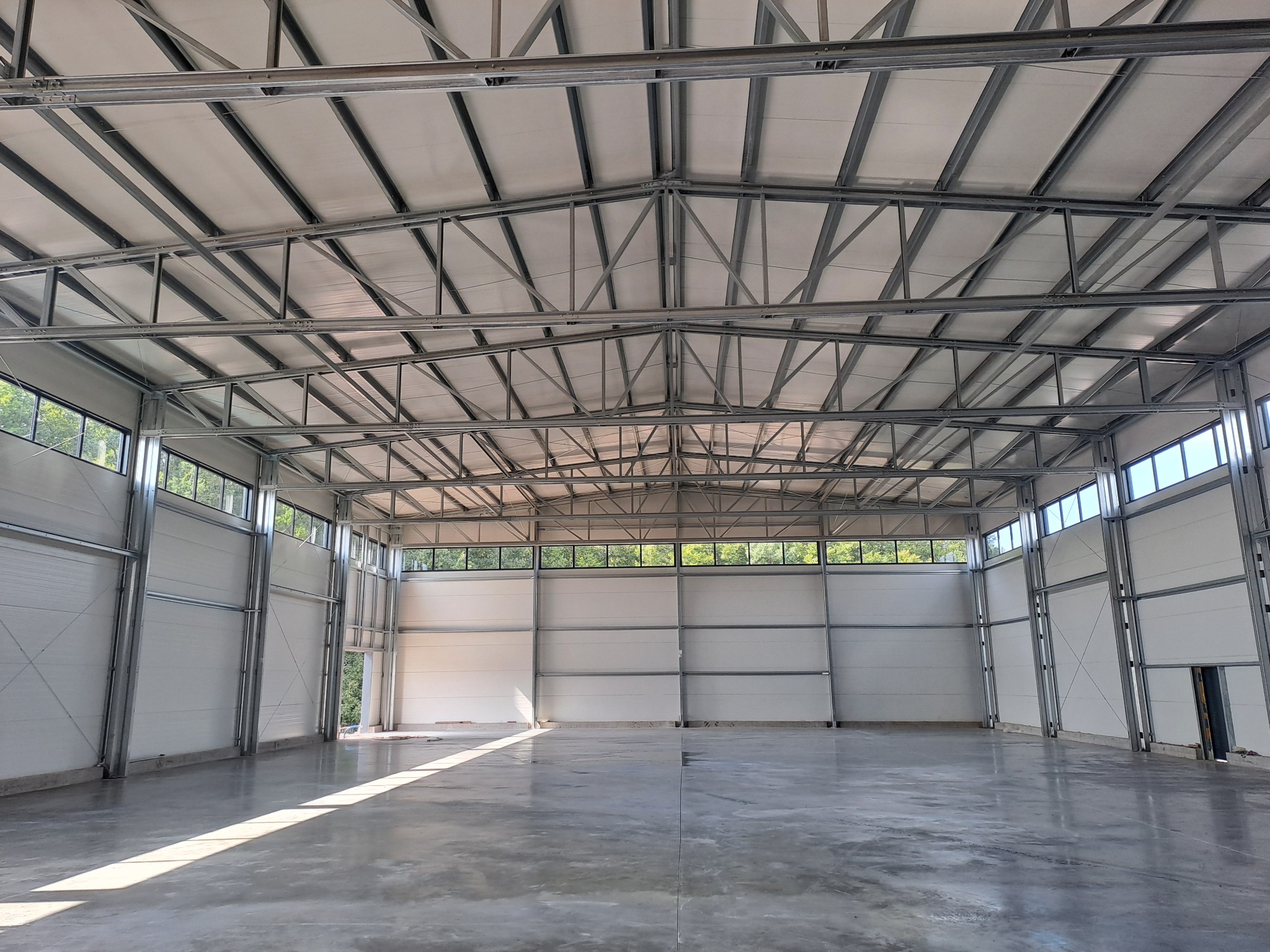 Wnętrze nowo wybudowanej hali z widoczną metalową konstrukcją dachu i ścianami z płyt warstwowych, betonowa posadzka z odbijającym się światłem słonecznym.