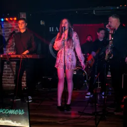 Wokalistka w cekinowej sukience śpiewa do mikrofonu na scenie z zespołem grającym na instrumentach, w tle logo klubu i czerwona kotara.