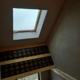 Montaż płyty karton gips na poddaszu ze skośnym sufitem, widoczne okno dachowe i częściowo otynkowana ściana.