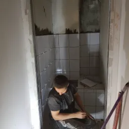 Układanie białych płytek w małej łazience podczas remontu, widoczne krzyżyki dystansowe, mężczyzna w ubrudzonym ubraniu roboczym, poziomica oparta o ścianę.