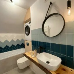 Nowoczesna łazienka na poddaszu z pralką obok toalety, umywalką na blacie z surowego drewna i wzorem chevron z płytek w odcieniach niebieskiego.