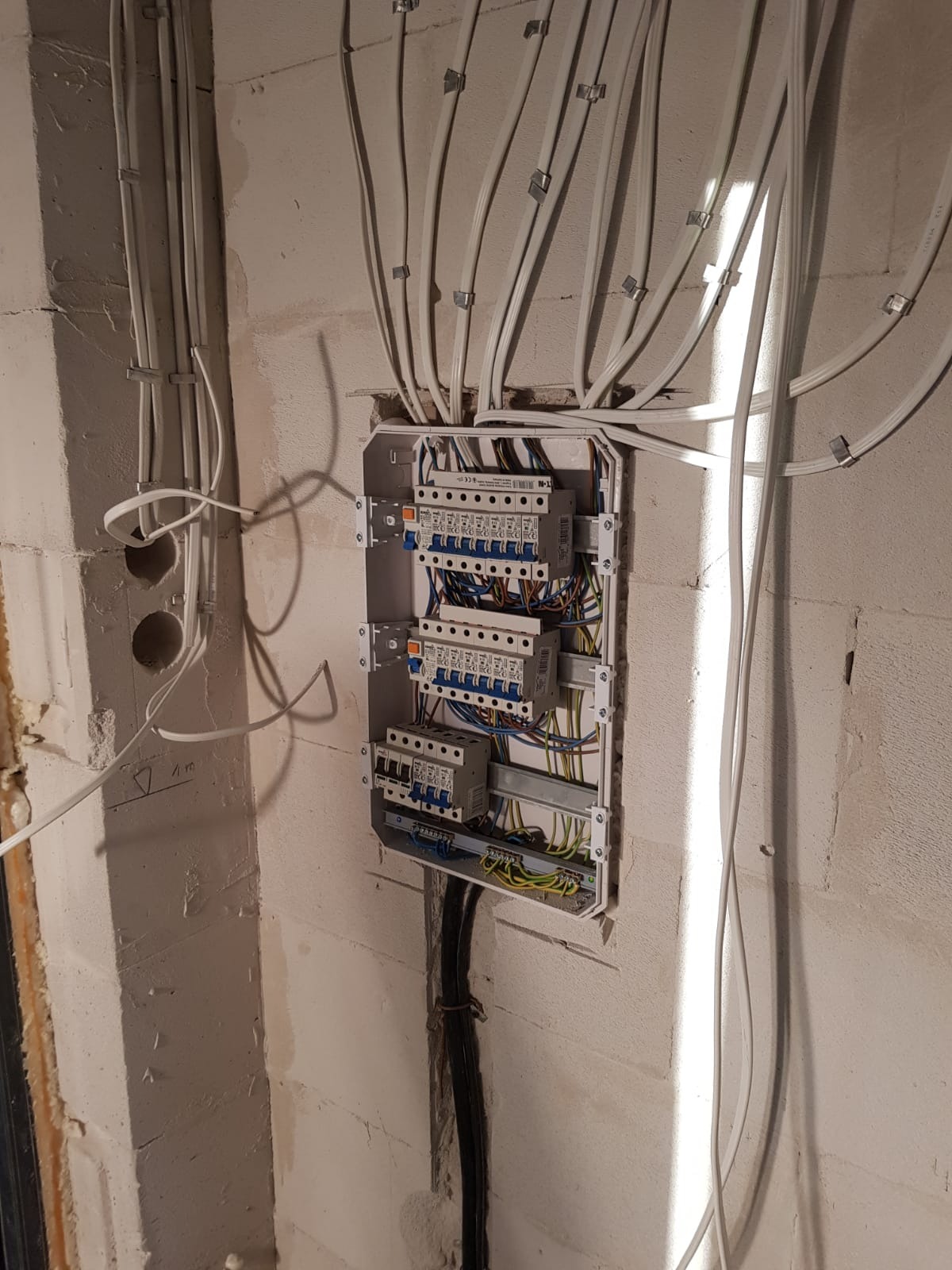 Rozdzielnica elektryczna z zamontowanymi bezpiecznikami i licznikiem, widoczne kable wchodzące i wychodzące ze ściany z surowego betonu, zabezpieczone metalowymi uchwytami.