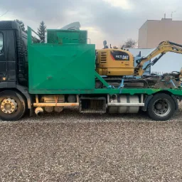 Żółta minikoparka CAT 302.7D CR zabezpieczona pasami na zielonej platformie transportowej ciężarówki Volvo, widok z boku na żwirowym podłożu.