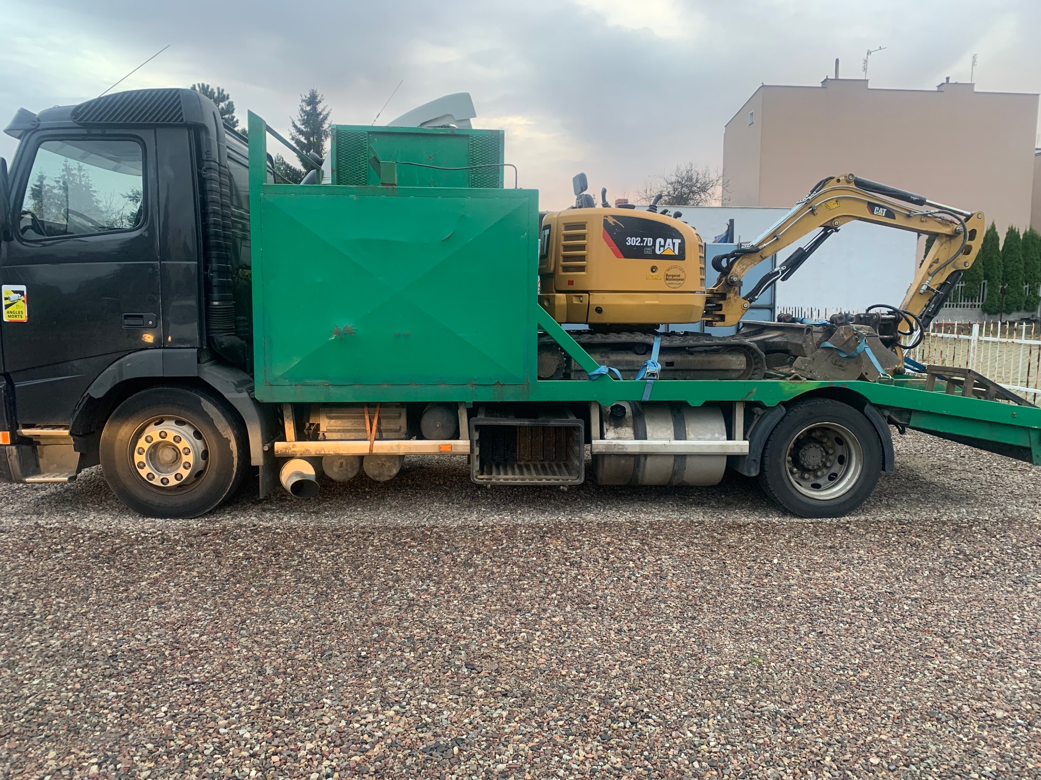 Żółta minikoparka CAT 302.7D CR zabezpieczona pasami na zielonej platformie transportowej ciężarówki Volvo, widok z boku na żwirowym podłożu.