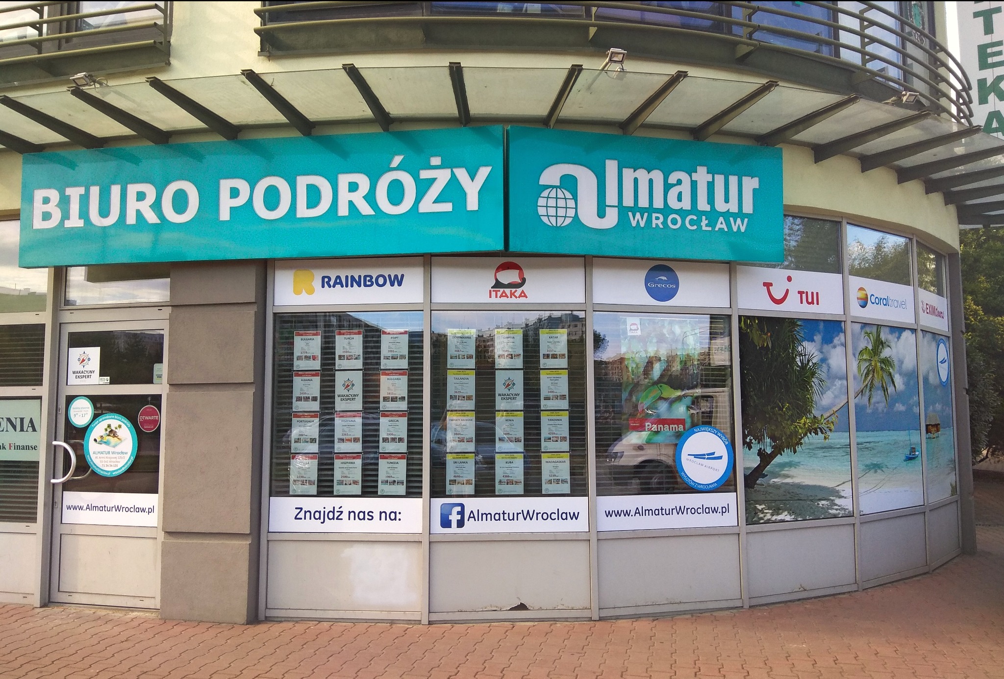 Fasada budynku z szyldem 'BIURO PODRÓŻY Almatur Wrocław', plakaty ofert wakacyjnych w oknach, logotypy Rainbow, Itaka, TUI i Coral Travel.