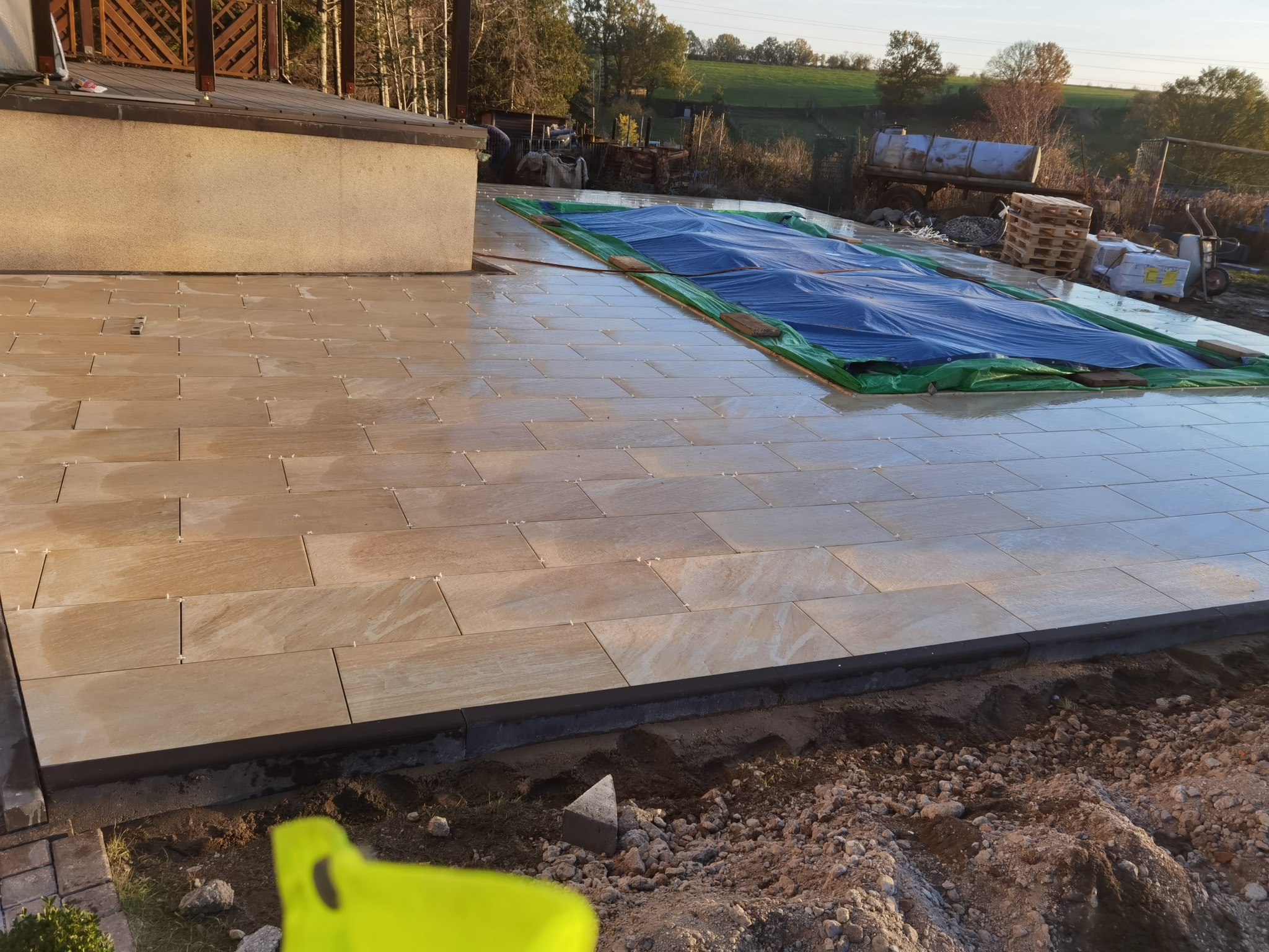 Układanie jasnobrązowej terakoty na tarasie, widoczne krzyżyki dystansowe, w tle przykryty basen i krajobraz wiejski.