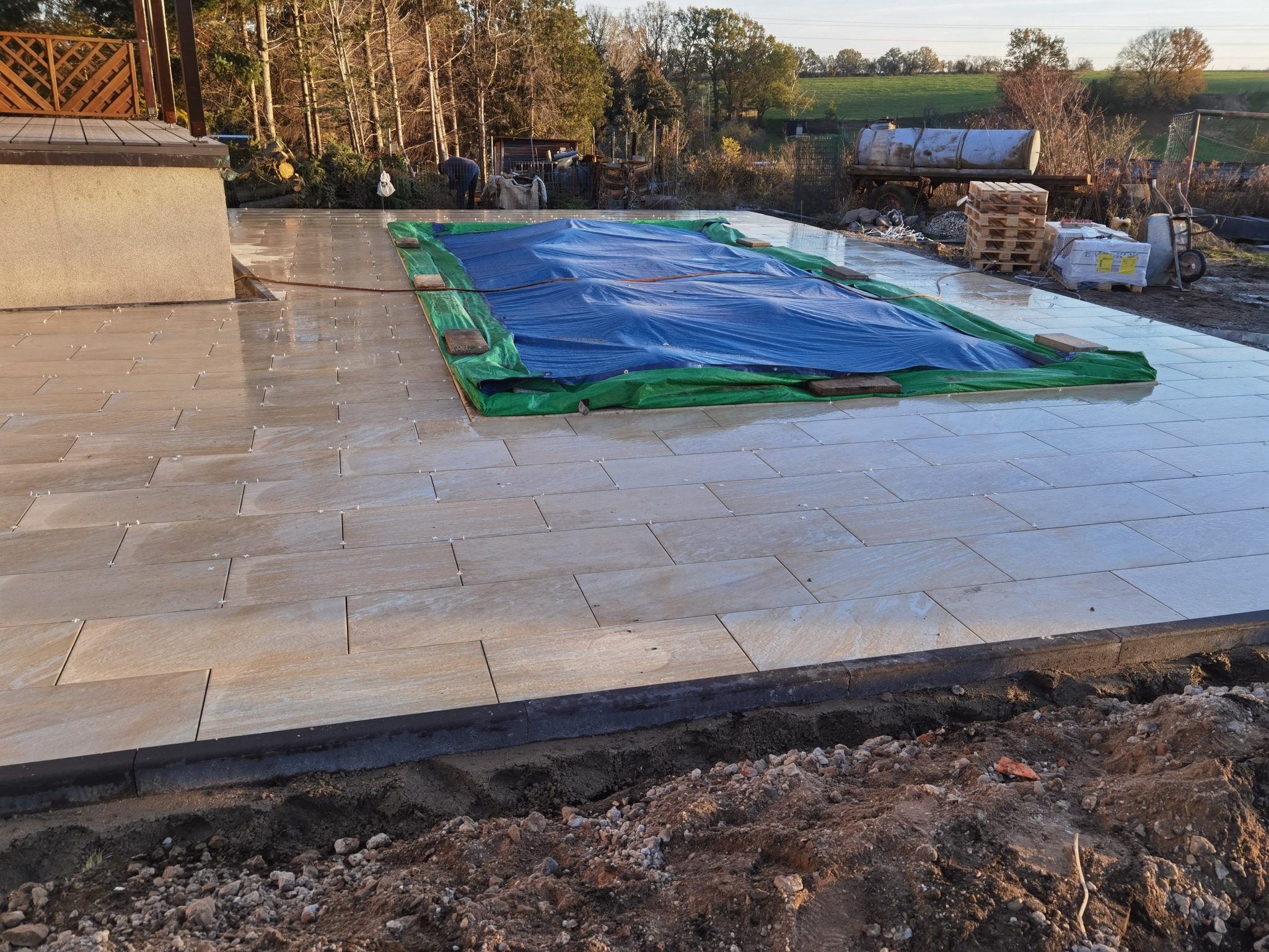 Układanie jasnych, prostokątnych płytek na tarasie w trakcie realizacji, widoczne plastikowe krzyżyki dystansowe, zabezpieczone plandeką miejsce na basen, w tle drzewa i zabudowania gospodarcze.