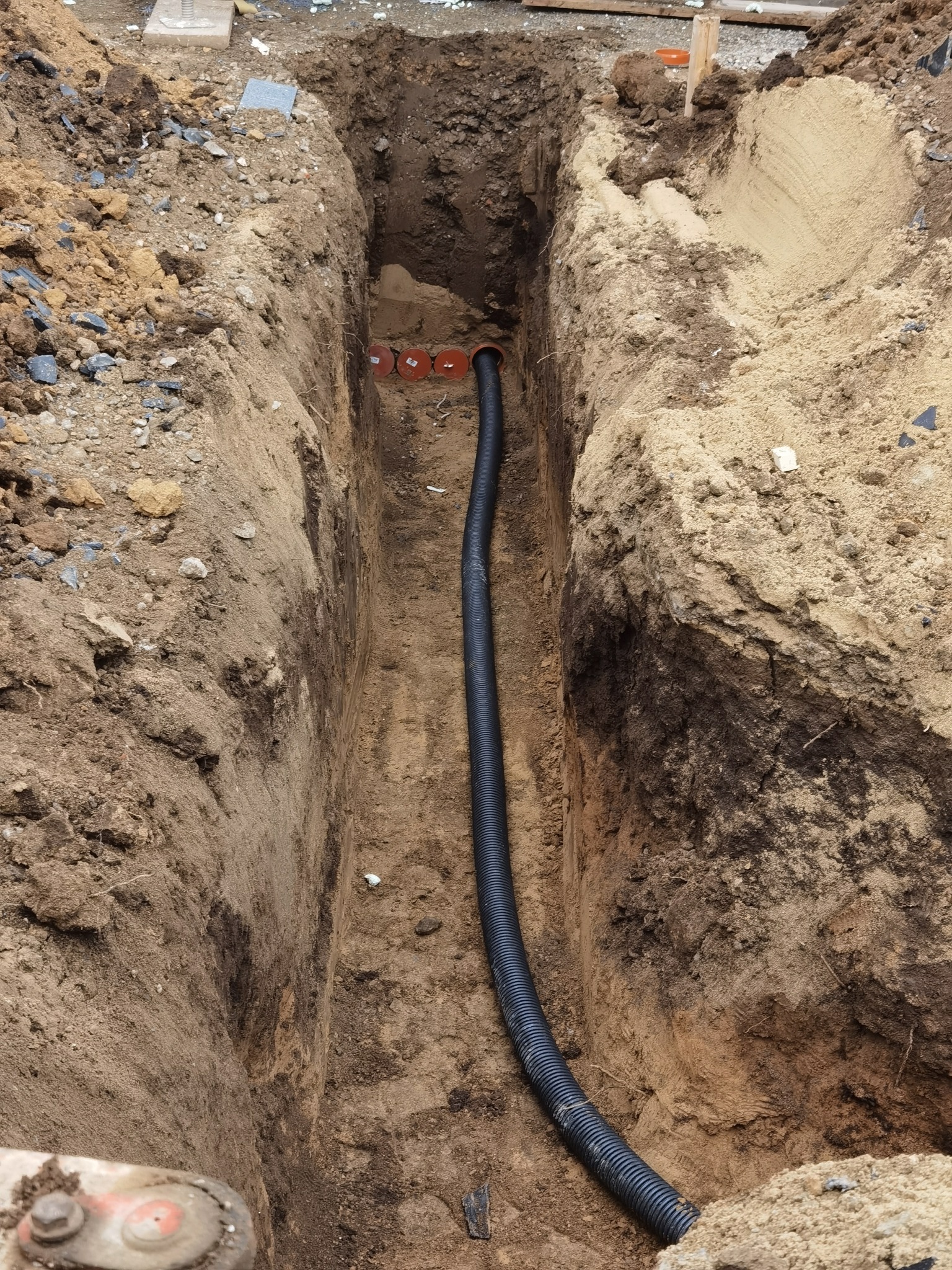 Wąski wykop w ziemi z ułożoną czarną rurą karbowaną i pomarańczowymi przepustami w tle.