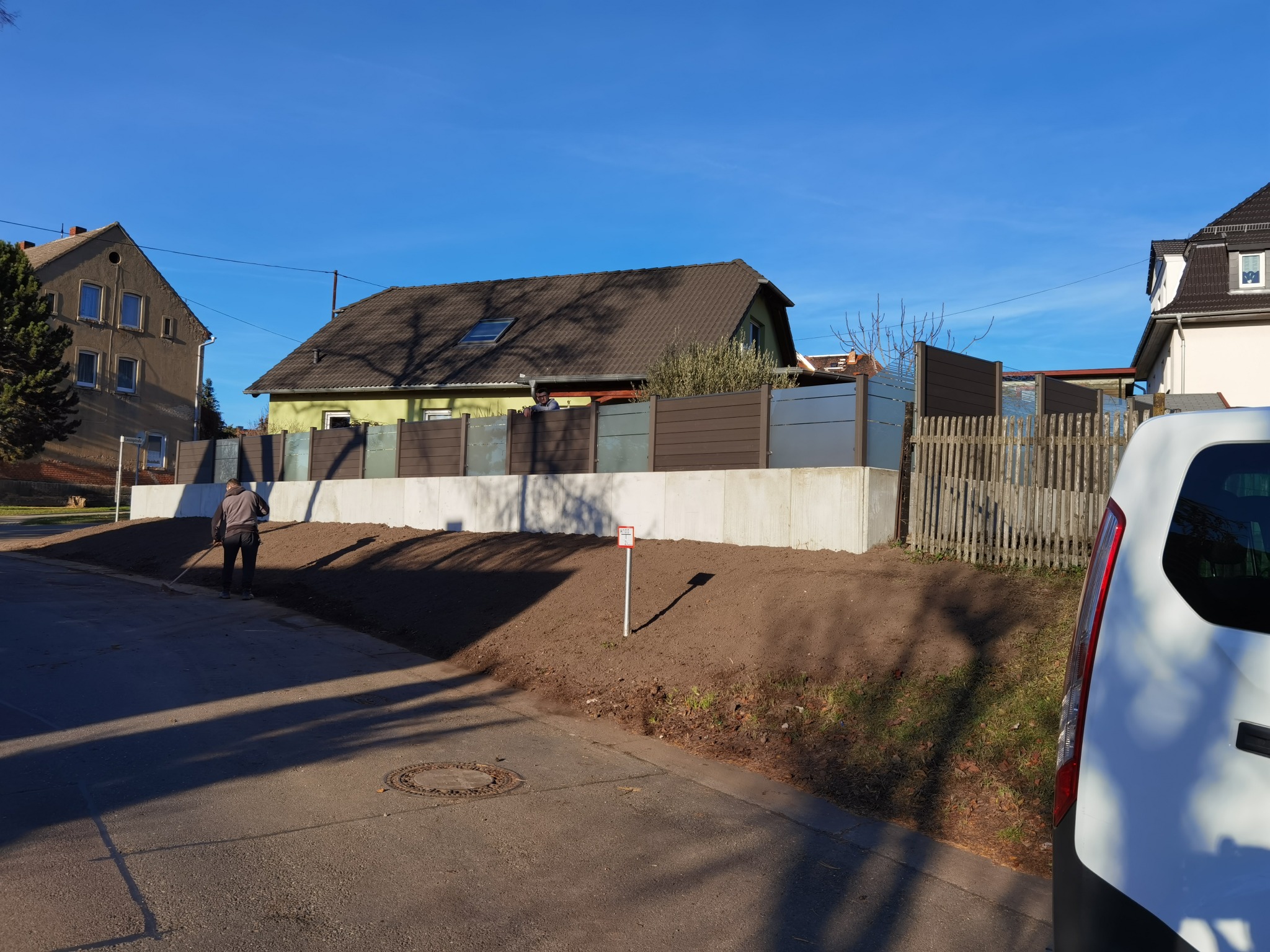 Nowoczesne ogrodzenie panelowe z elementami przezroczystego szkła, zamontowane na betonowej podmurówce, z pracownikiem wyrównującym teren wokół, na tle zabudowy jednorodzinnej w słoneczny dzień.