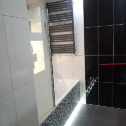 Nowoczesna łazienka z czarno-białymi płytkami, podświetlaną wanną obudowaną mozaiką i grzejnikiem drabinkowym, widok z boku.