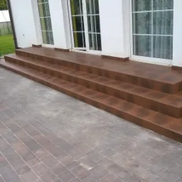 Zewnętrzne schody wejściowe do domu wyłożone brązowymi płytkami ceramicznymi, z widoczną kostką brukową na podwórku i fragmentem ogrodu z zielenią w tle.